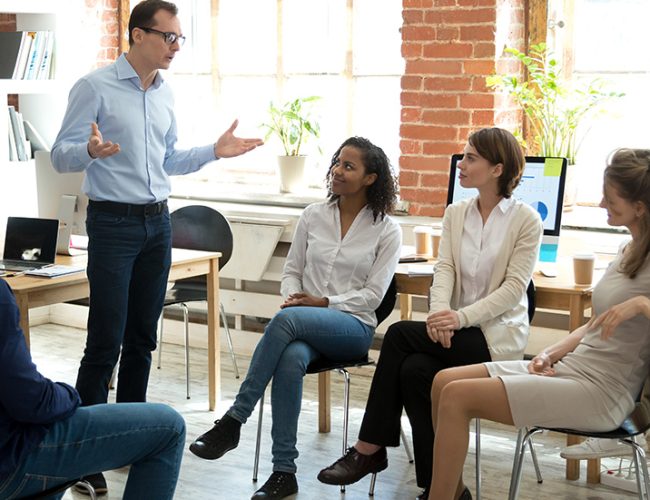 Diverse business team employees listening to male manager coach speaking at group office meeting, mentor executive leader talking during briefing, multi-ethnic workers engaged in corporate training